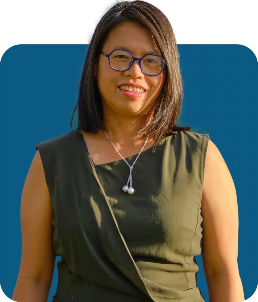 A smiling woman with shoulder-length dark brown hair, wearing glasses, a sleeveless olive green top, and a pearl necklace, standing against a blue background.