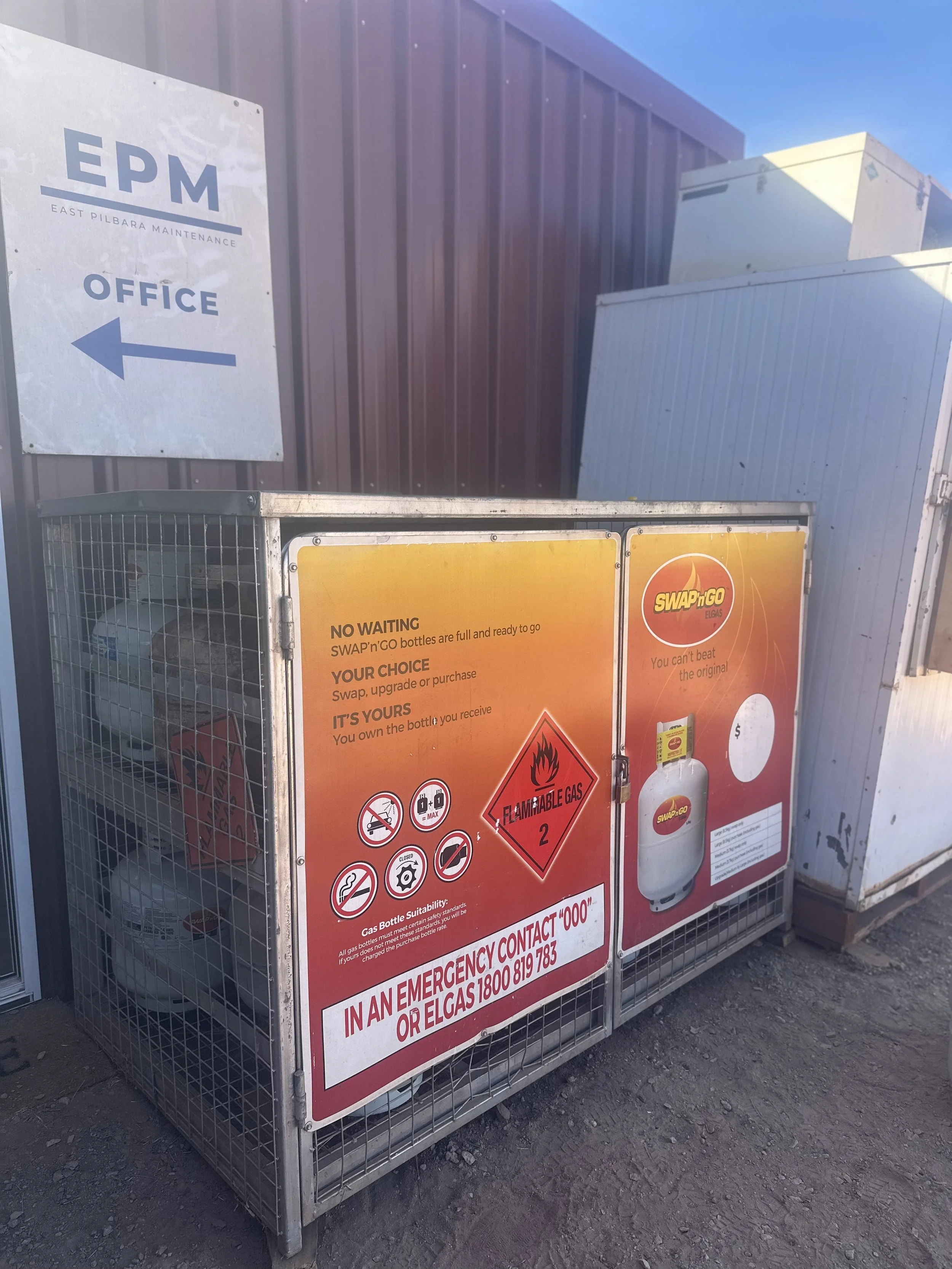 A metal cage containing gas bottles labeled 'SWAPn'GO ELGAS,' with warning signs, including a flammable gas warning, and instructions for emergencies. Behind the cage, there is a purple building with a sign pointing to the office, and the ground is dirt.
