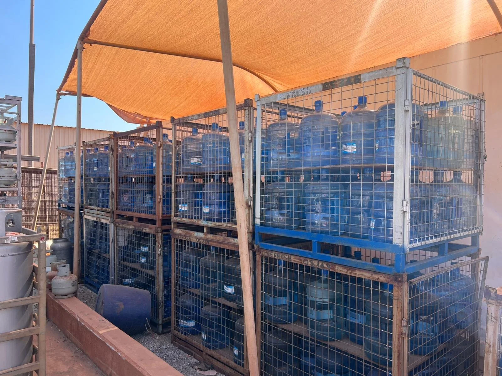 Stacked large water bottles in metal cages outdoors under an orange canopy.