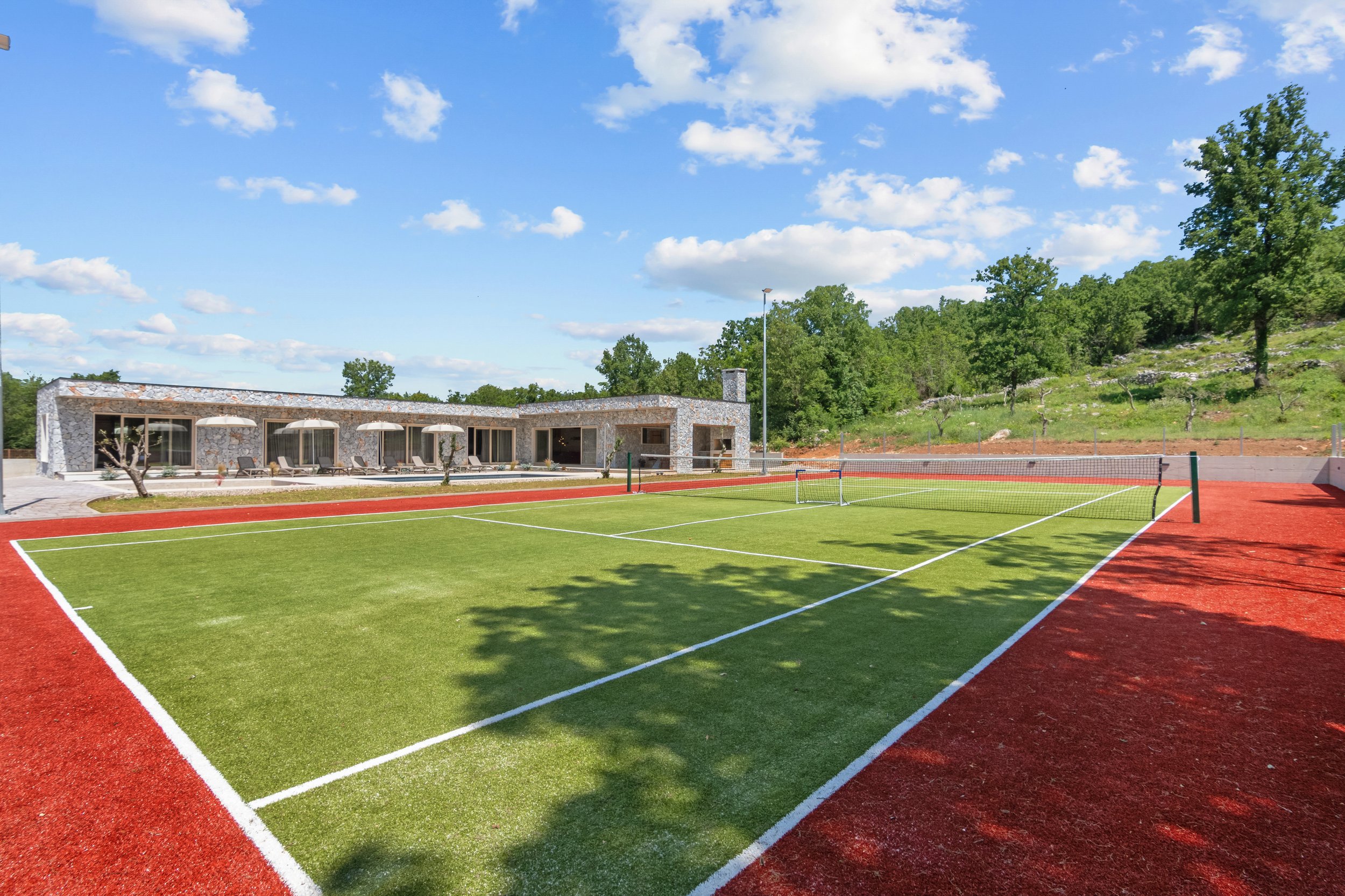 An outdoor tennis court with a green playing surface and red surrounding area, with a modern stone building and trees in the background under a blue sky with white clouds.