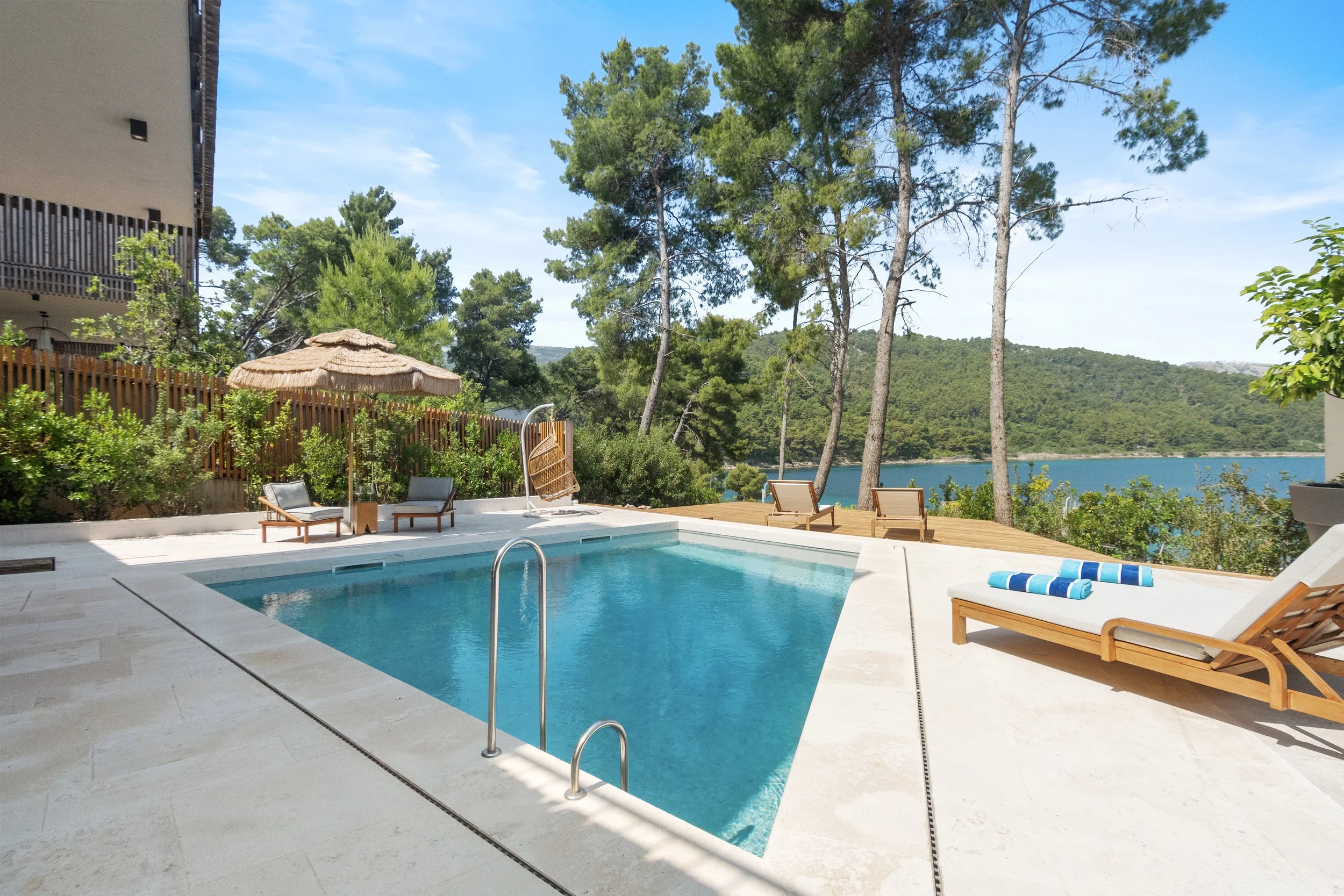 Swimming pool area with lounge chairs, a thatched umbrella, and a view of trees and water in the background under a clear sky.