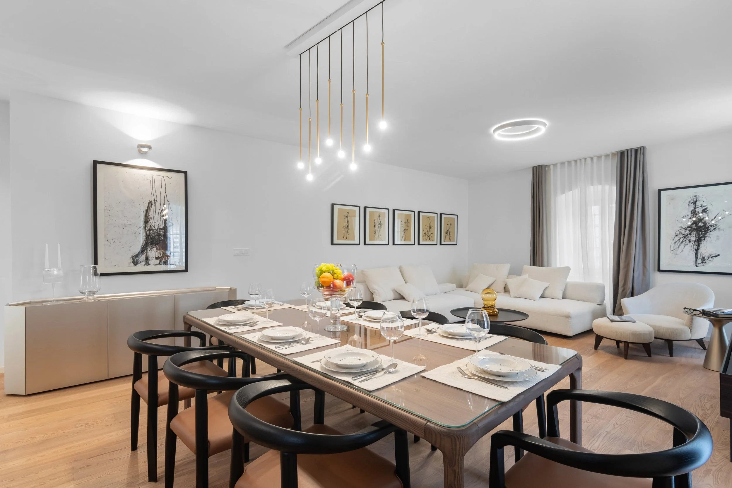 Modern dining room with a wooden table set for a meal, surrounded by black and tan chairs, with a white sofa and artwork in the background.