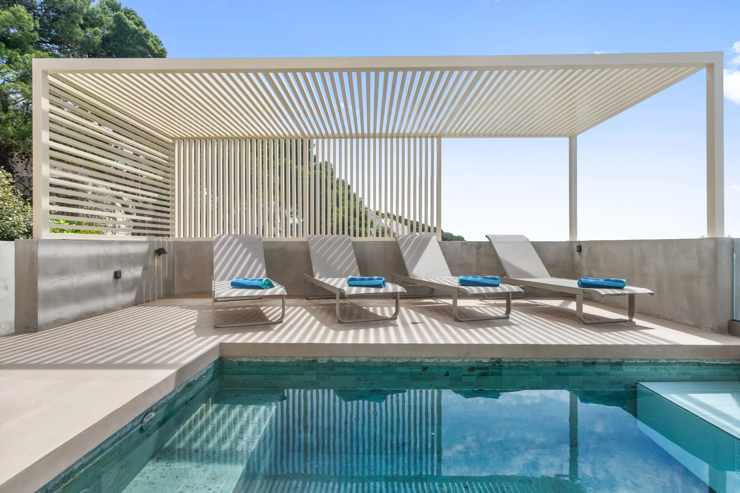 Poolside lounge chairs with folded towels under a shaded pergola near a swimming pool.