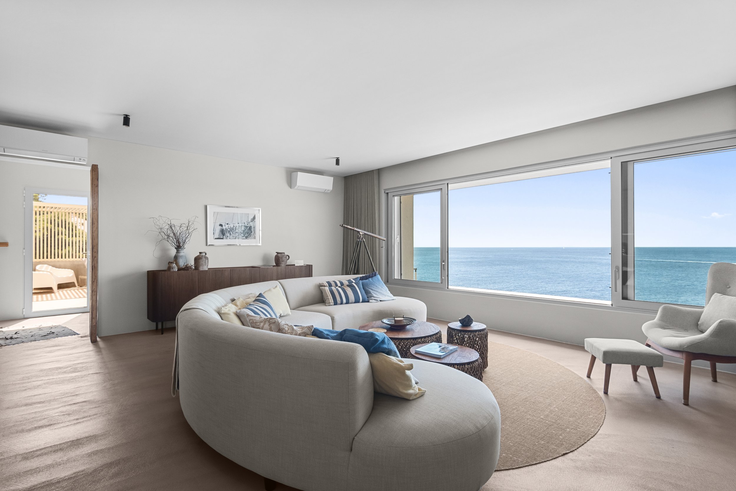 Living room with large windows overlooking the ocean, white curved sofa with striped pillows, armchair with footstool, wooden side tables, and ocean view.