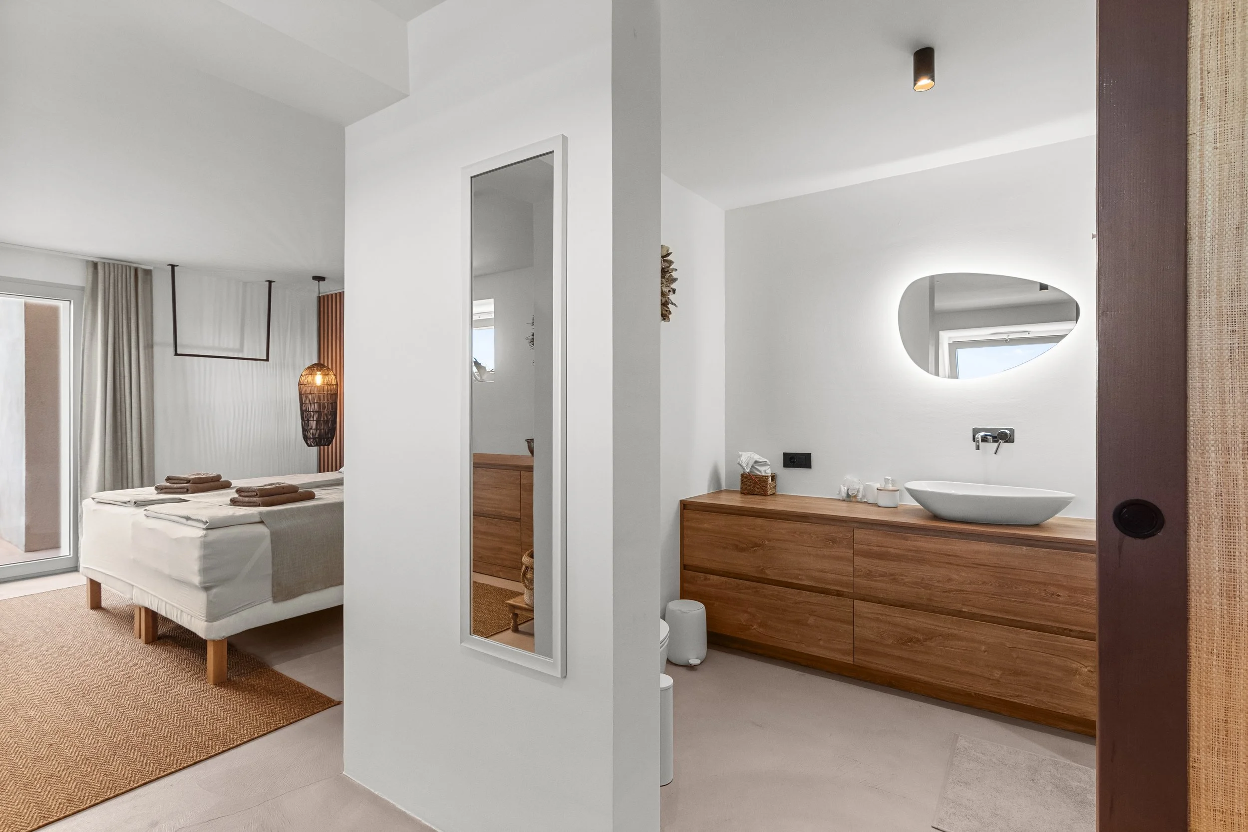 Bedroom with a balcony door and a bed made with white bedding and brown towels, adjacent to a bathroom with a wooden vanity, oval mirror with backlighting, and a vessel sink.