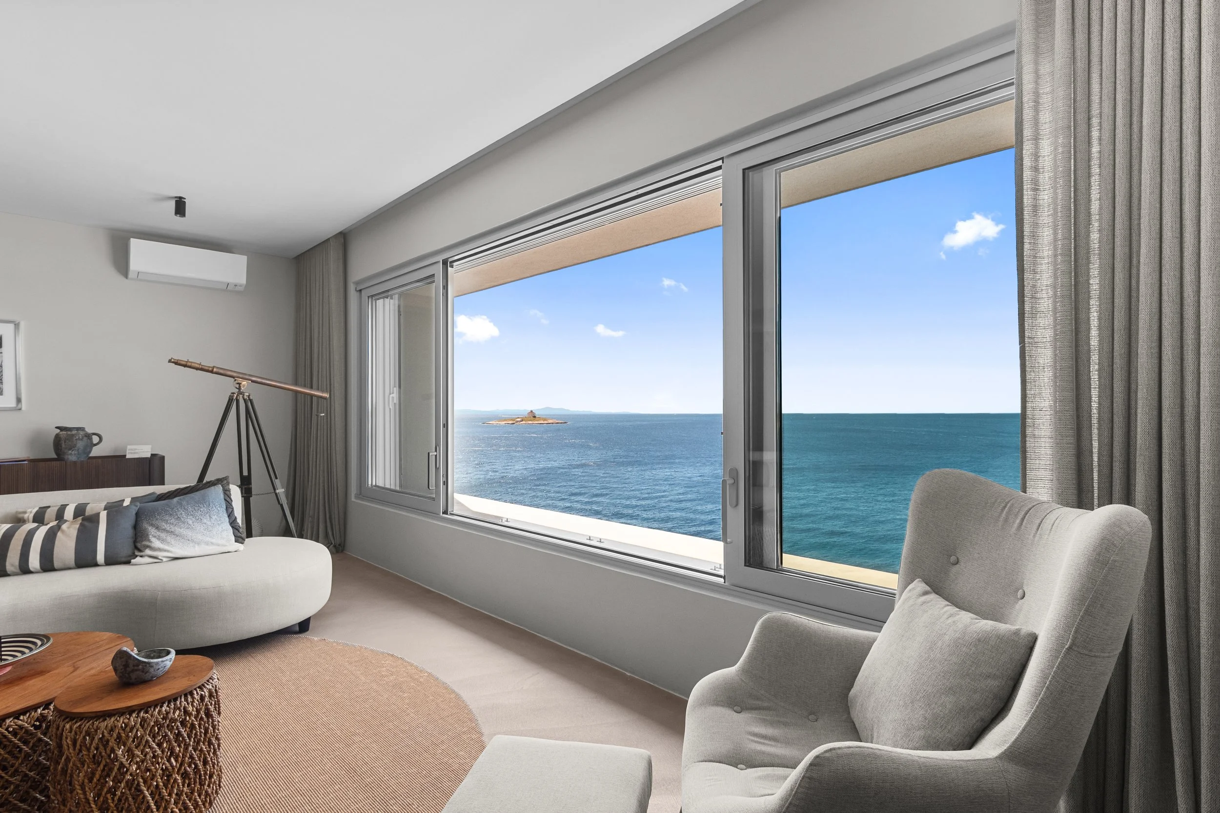 Living room with large window overlooking ocean and clear blue sky, light-colored furniture, beige rug, telescope, and decorative items.