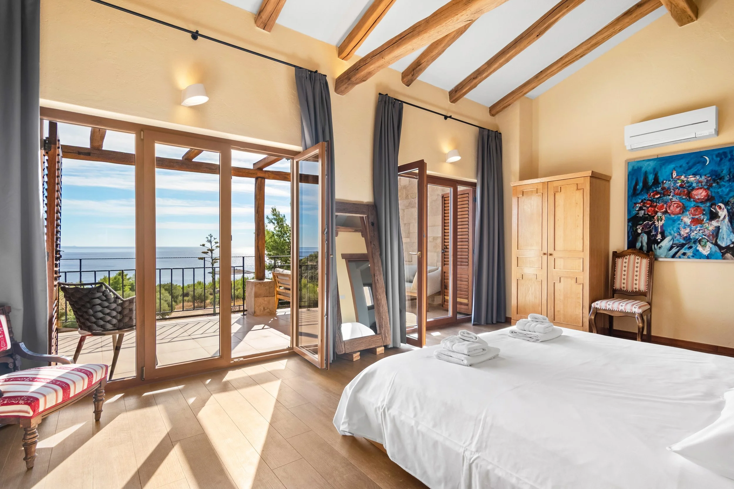 A cozy villa room with a large bed, wooden beams on the ceiling, and a balcony with a view of the ocean and greenery.