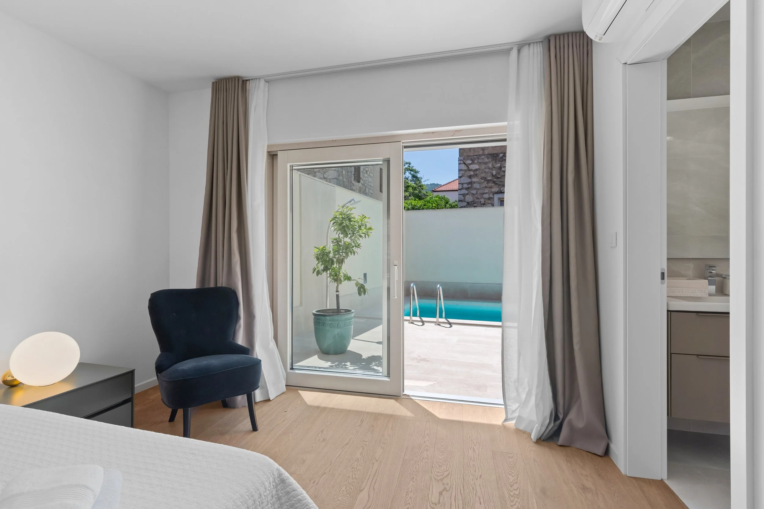 A bedroom with a sliding glass door leading to an outdoor patio and swimming pool, white walls, a blue armchair, a bedside table with a modern lamp, and part of a bathroom visible on the right.