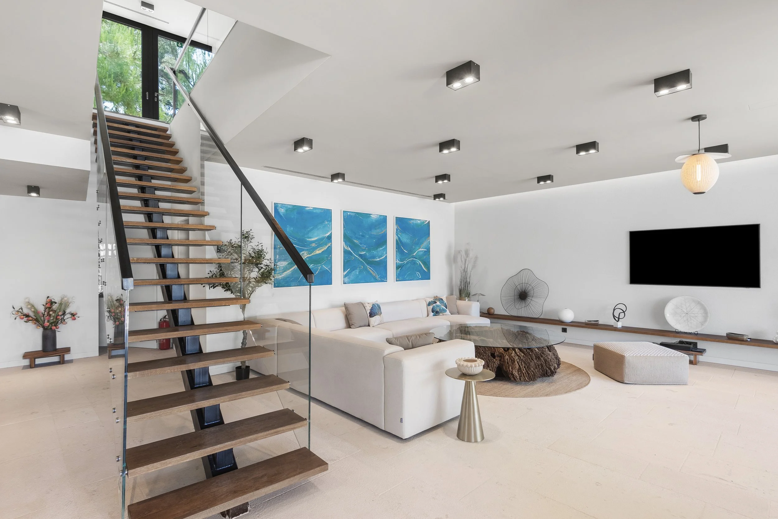 Modern living room with white L-shaped sofa, blue abstract artwork on wall, glass-top coffee table with textured base, flat-screen TV, wall decor, plants, and a open staircase with wooden steps and black railing.