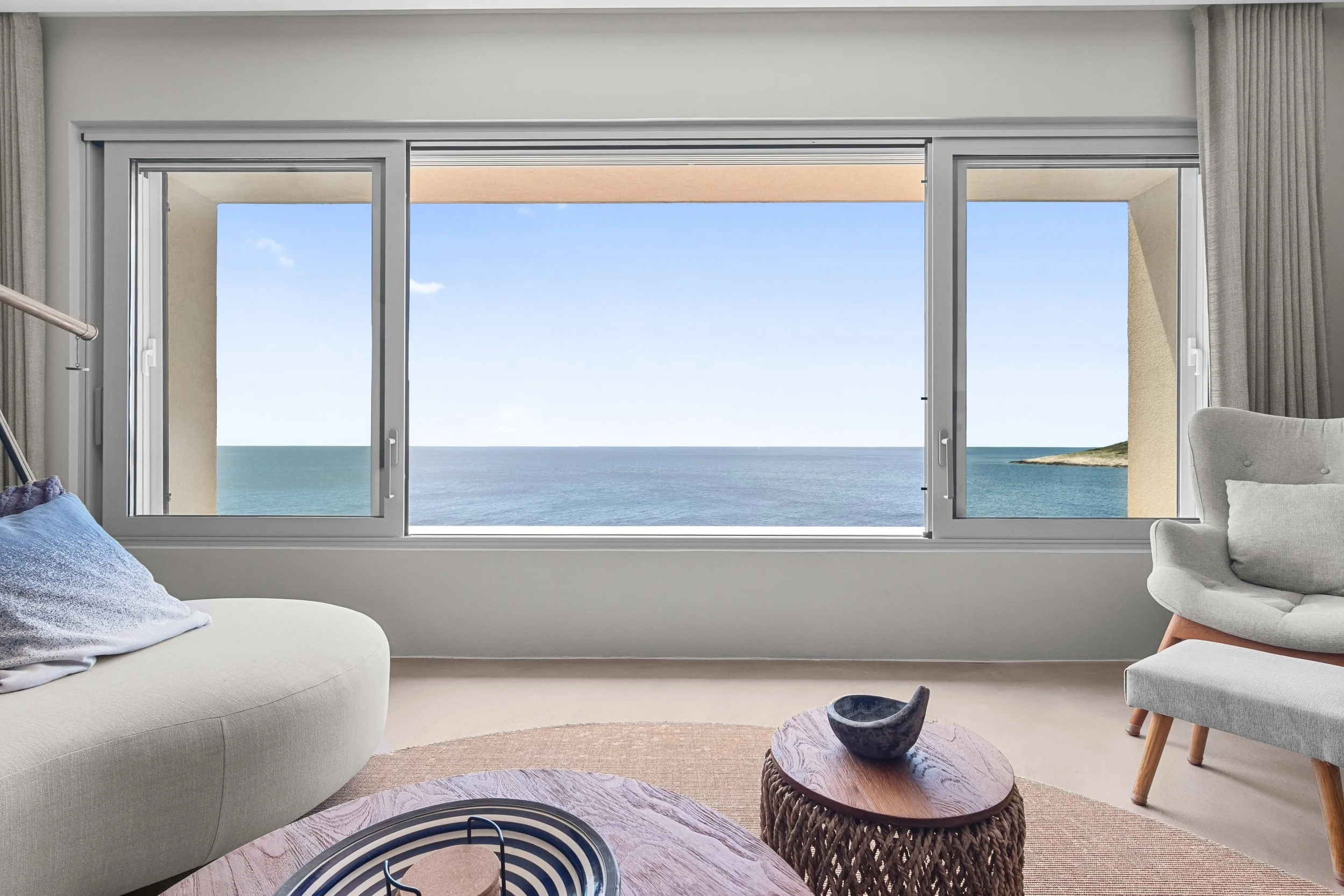 Living room with large window view of the ocean and blue sky, beige couch and gray armchair.
