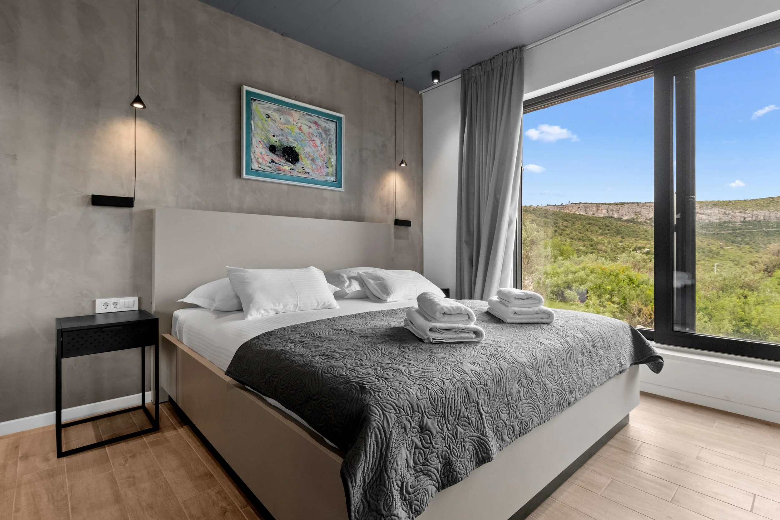 Modern bedroom with a large bed, black quilt, and white pillows, next to a floor-to-ceiling window showing green hills and blue sky, with artwork above the bed and a small black bedside table.