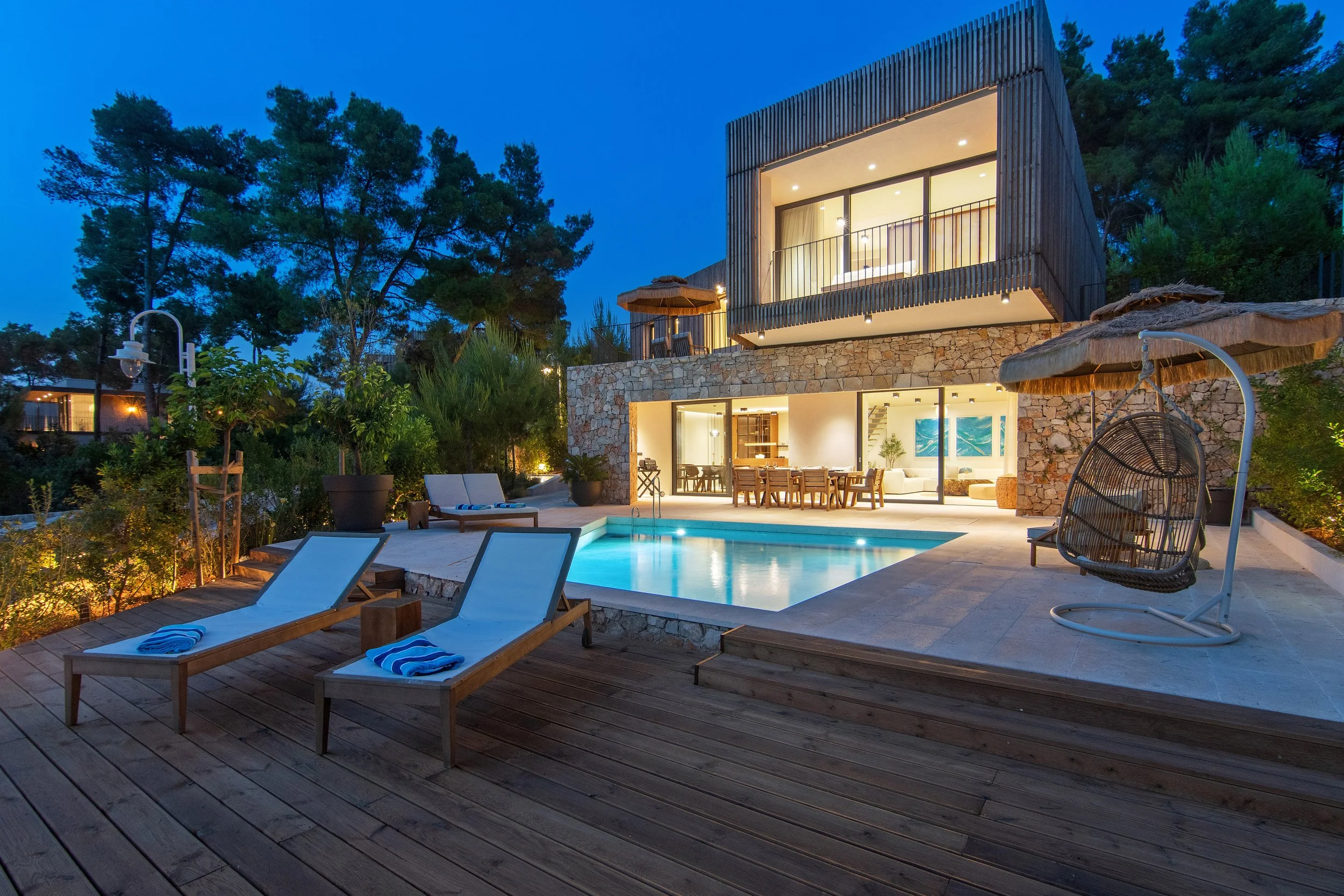 Luxury modern house with swimming pool, outdoor lounge chairs, and a hanging egg chair, surrounded by trees during evening.