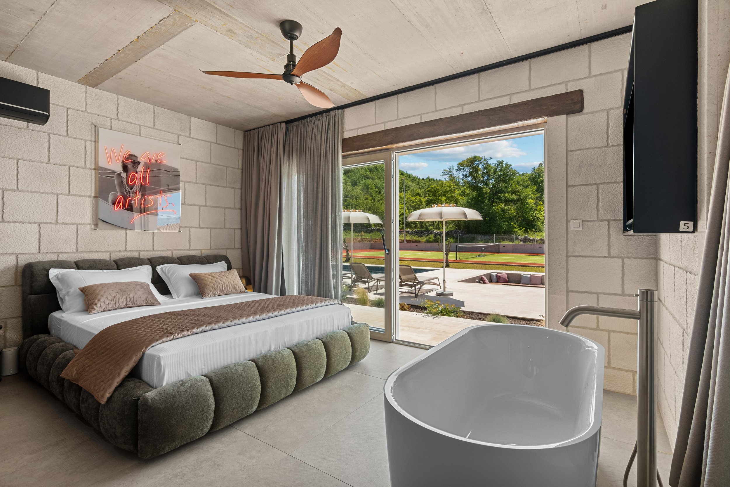 Modern bedroom with a large bed, neon art sign, sliding glass door opening to outdoor lounge chairs and umbrellas, and a freestanding bathtub.