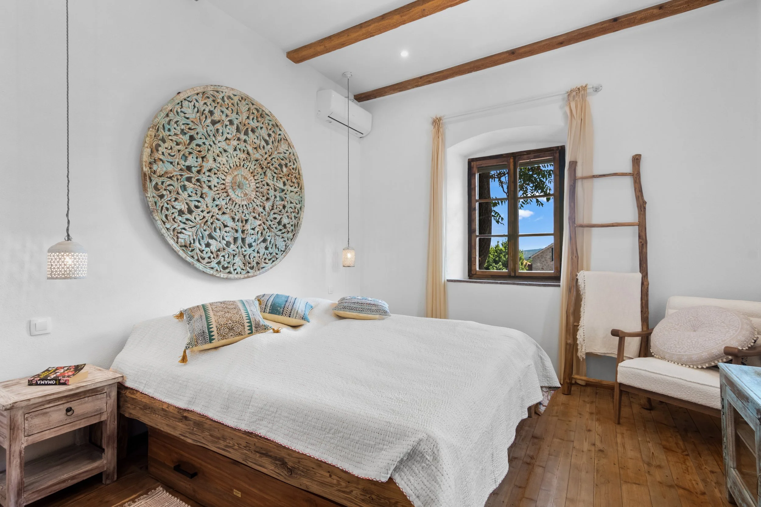 A bedroom with a white bed, rustic wooden furniture, a window with beige curtains, and decorative pillows. There is a large circular carved wall art above the bed, a hanging pendant light, a wooden ladder, and an outdoor view of blue sky and trees th