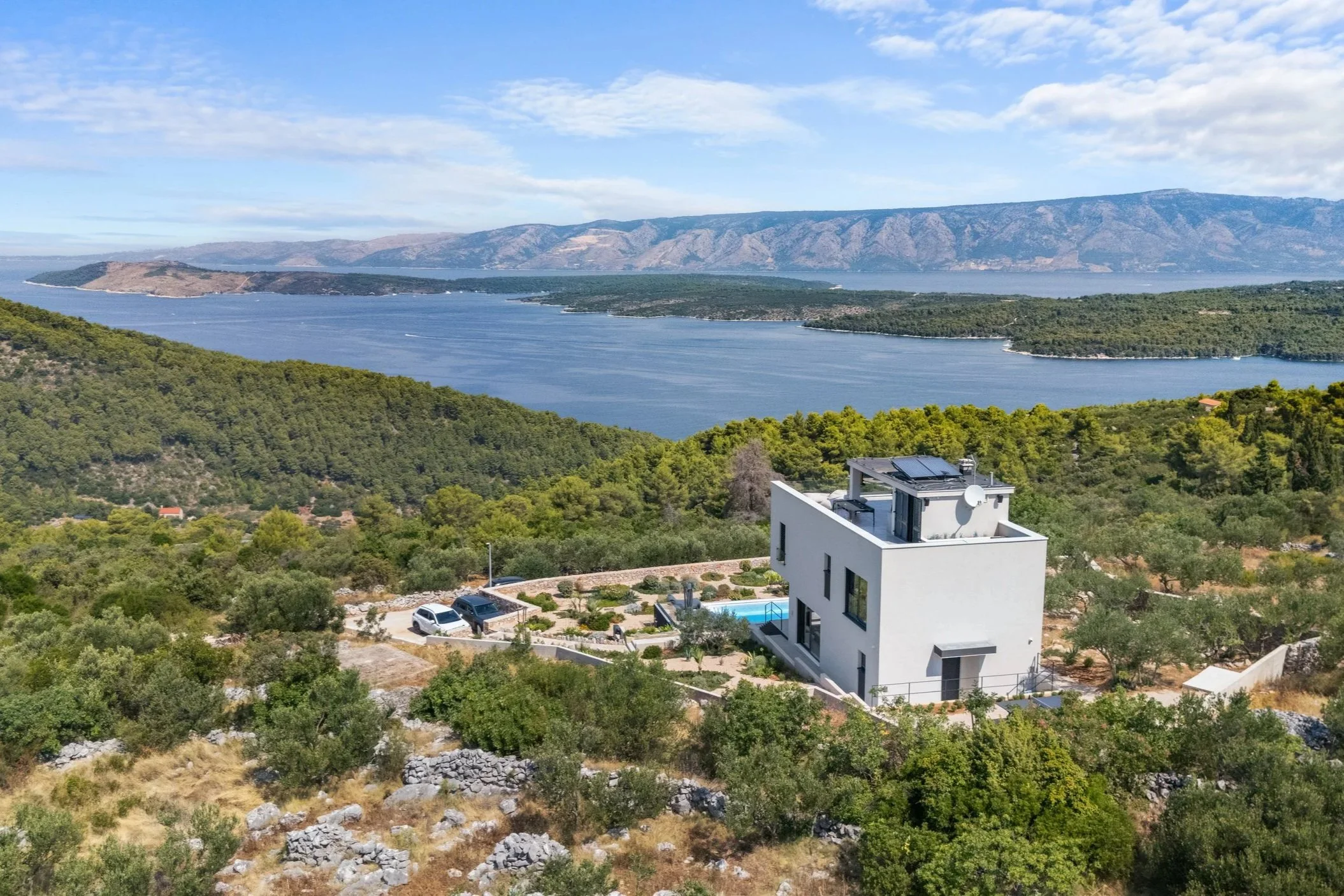Night view of a modern house with a lit swimming pool in the foreground, an outdoor patio with furniture, and large windows revealing the interior, with trees and a dark sky in the background.