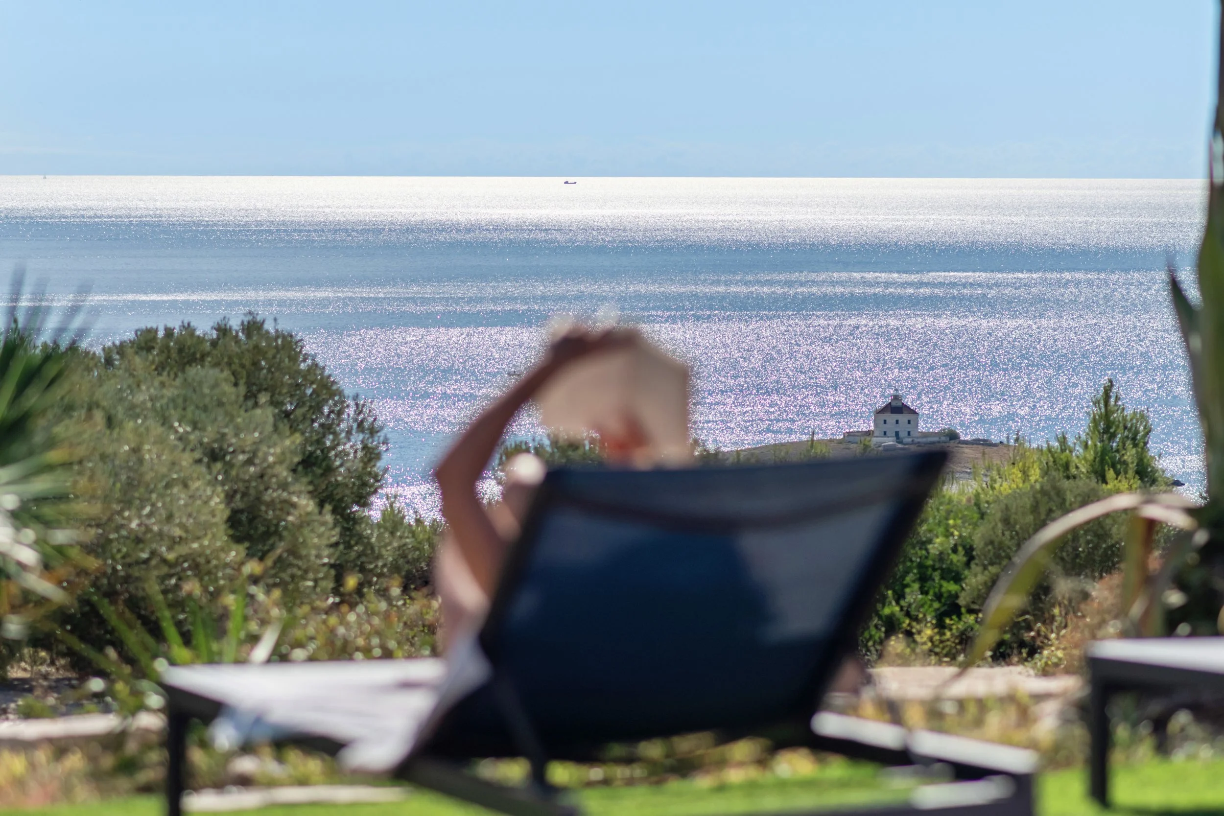 Person relaxing on a lounge chair at a seaside location, with a view of sparkling ocean and a lighthouse in the distance under a clear blue sky.