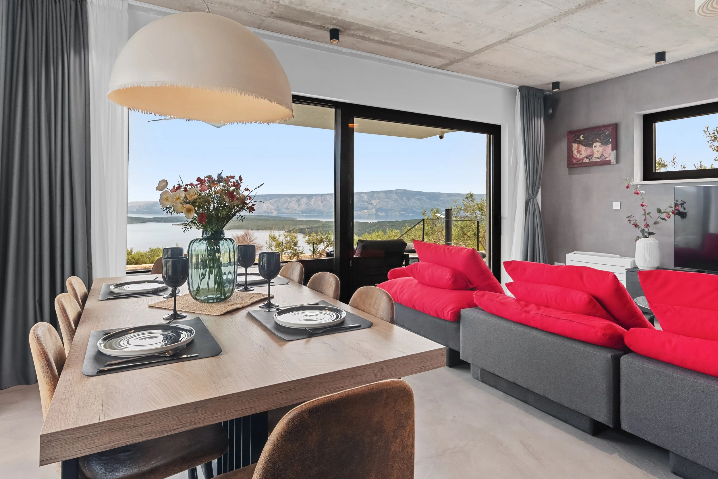 Modern living and dining room with a large sliding glass door opening to a lake view, grey and red color scheme, and minimalist decor.