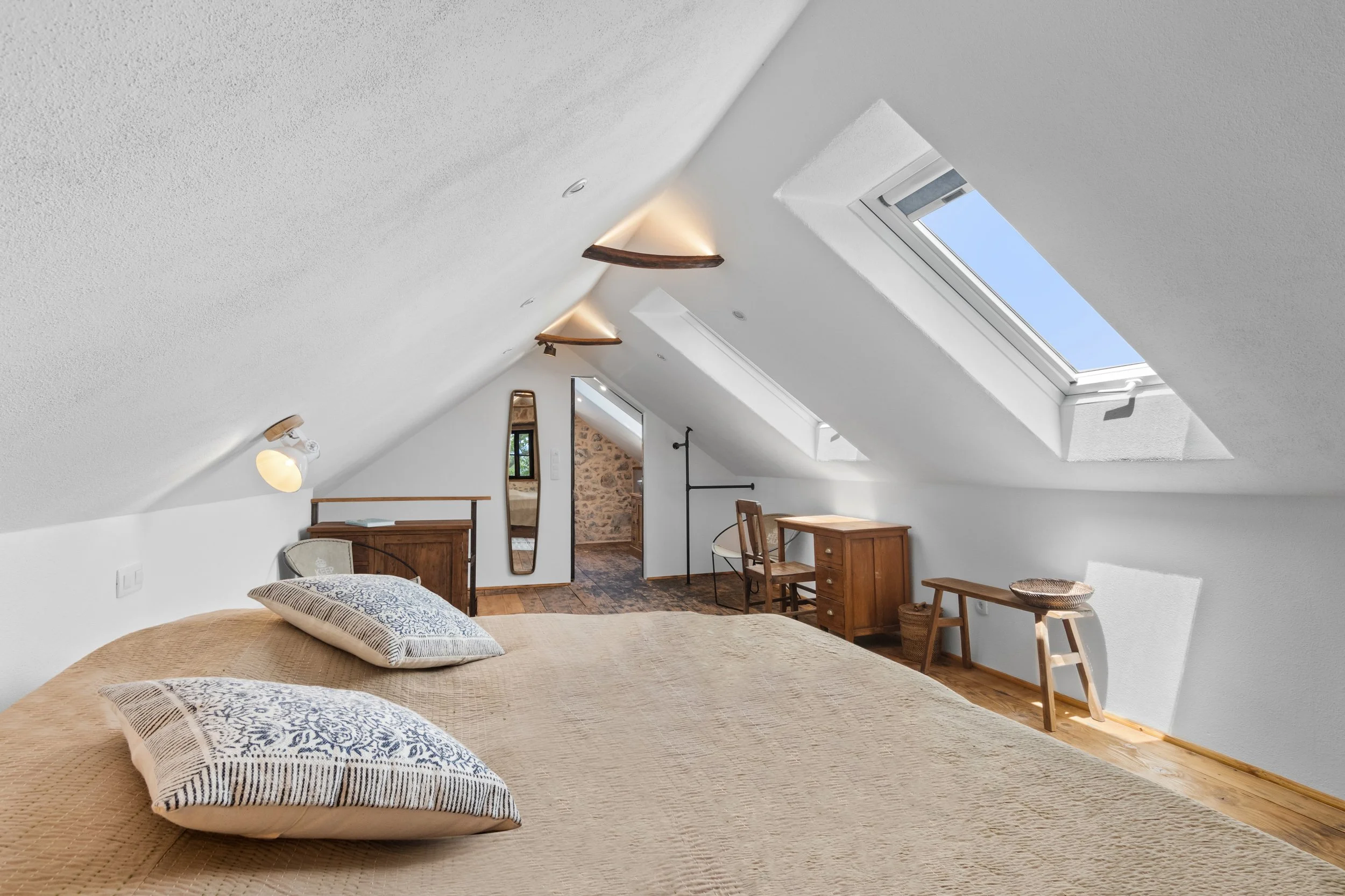 Bedroom with sloped ceiling and large skylight windows, wooden furniture including a desk and chairs, a bed with throw pillows, and a full-length mirror.
