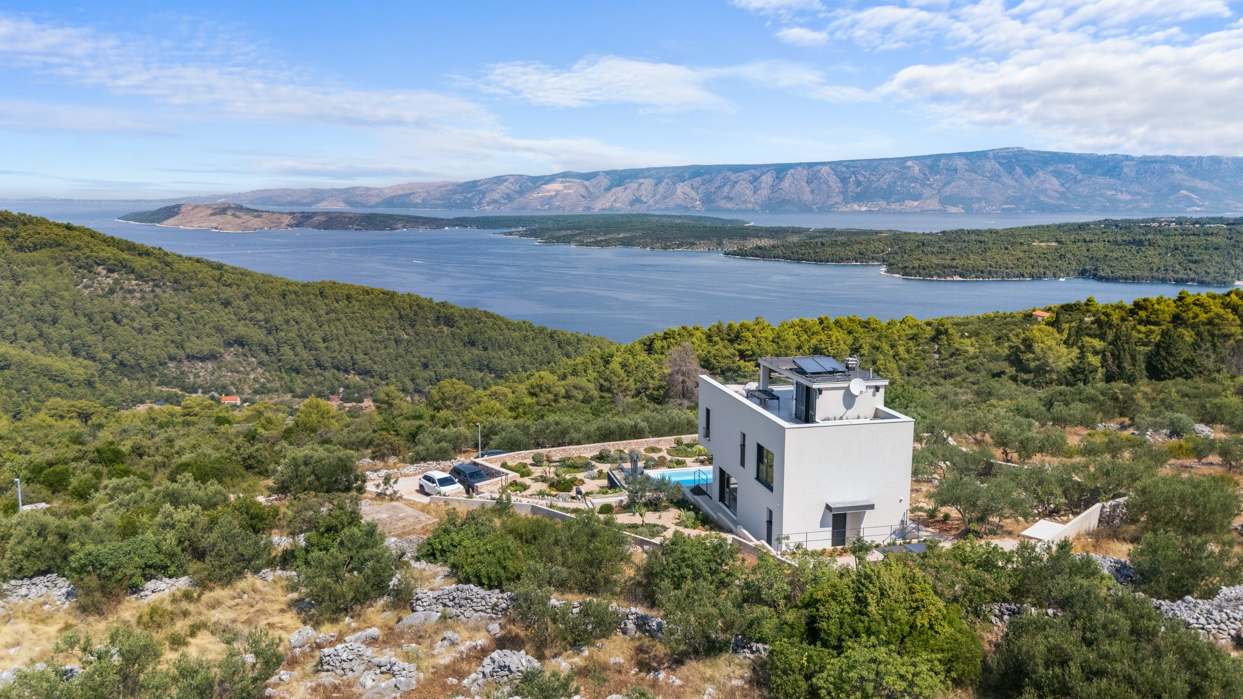 Modern white house with solar panels on the roof, situated on a hillside surrounded by trees and shrubs, overlooking a large body of water with mountains in the distance under a partly cloudy sky.