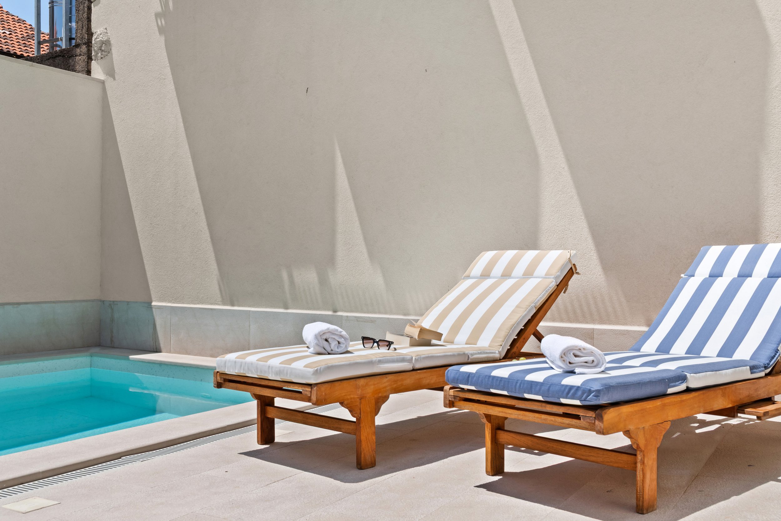 Two wooden poolside lounge chairs with striped cushions, one beige and white, the other blue and white, placed next to a small pool. Towels and sunglasses rest on the chairs. Shadows cast on a beige wall in the background.