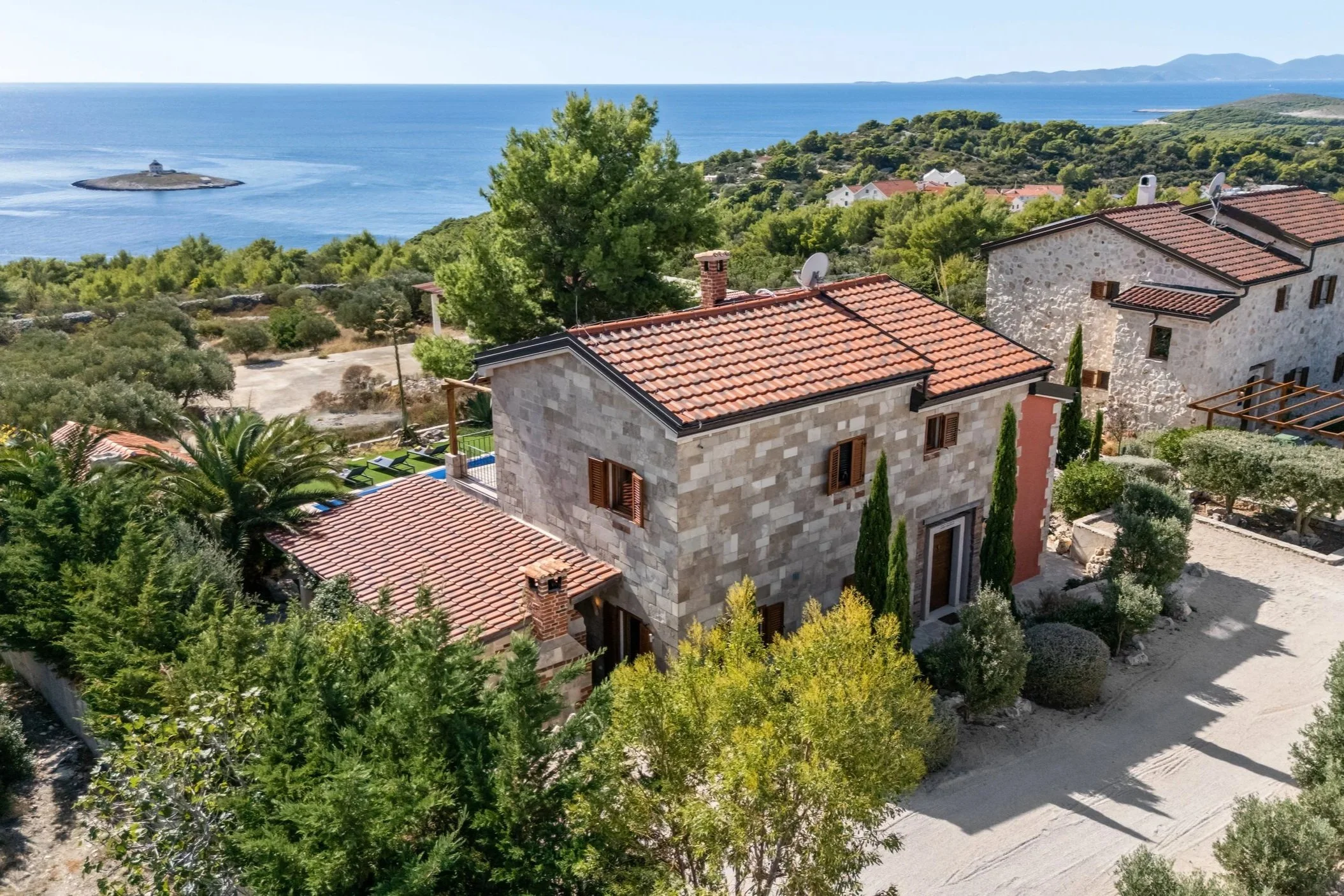Aerial view of a stone house with red tile roof, surrounded by green trees and shrubs, overlooking the coastline with a small island and water in the background.