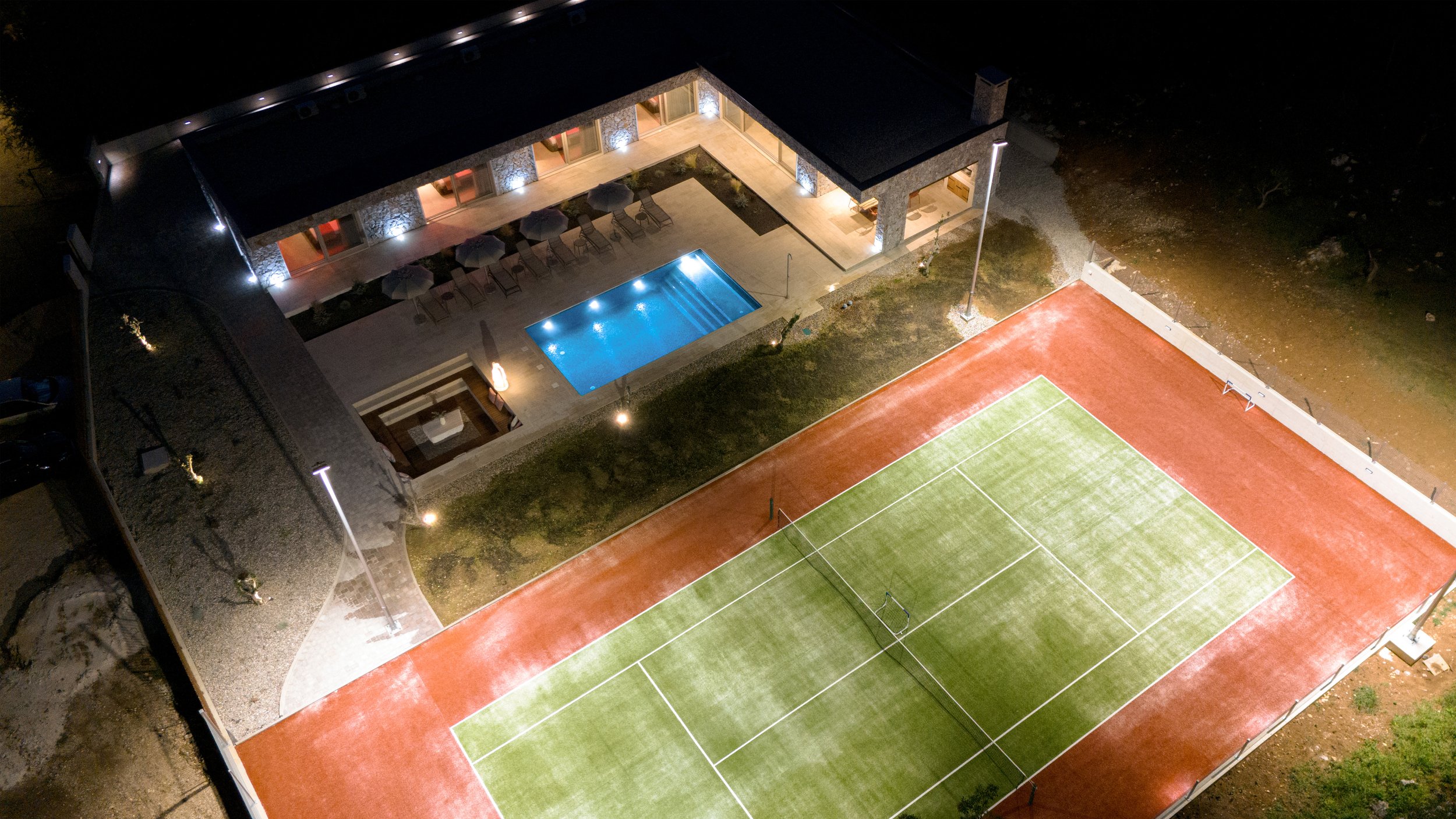 Night aerial view of a sports complex with a tennis court and swimming pool next to a building with patio seating, illuminated by exterior lights.