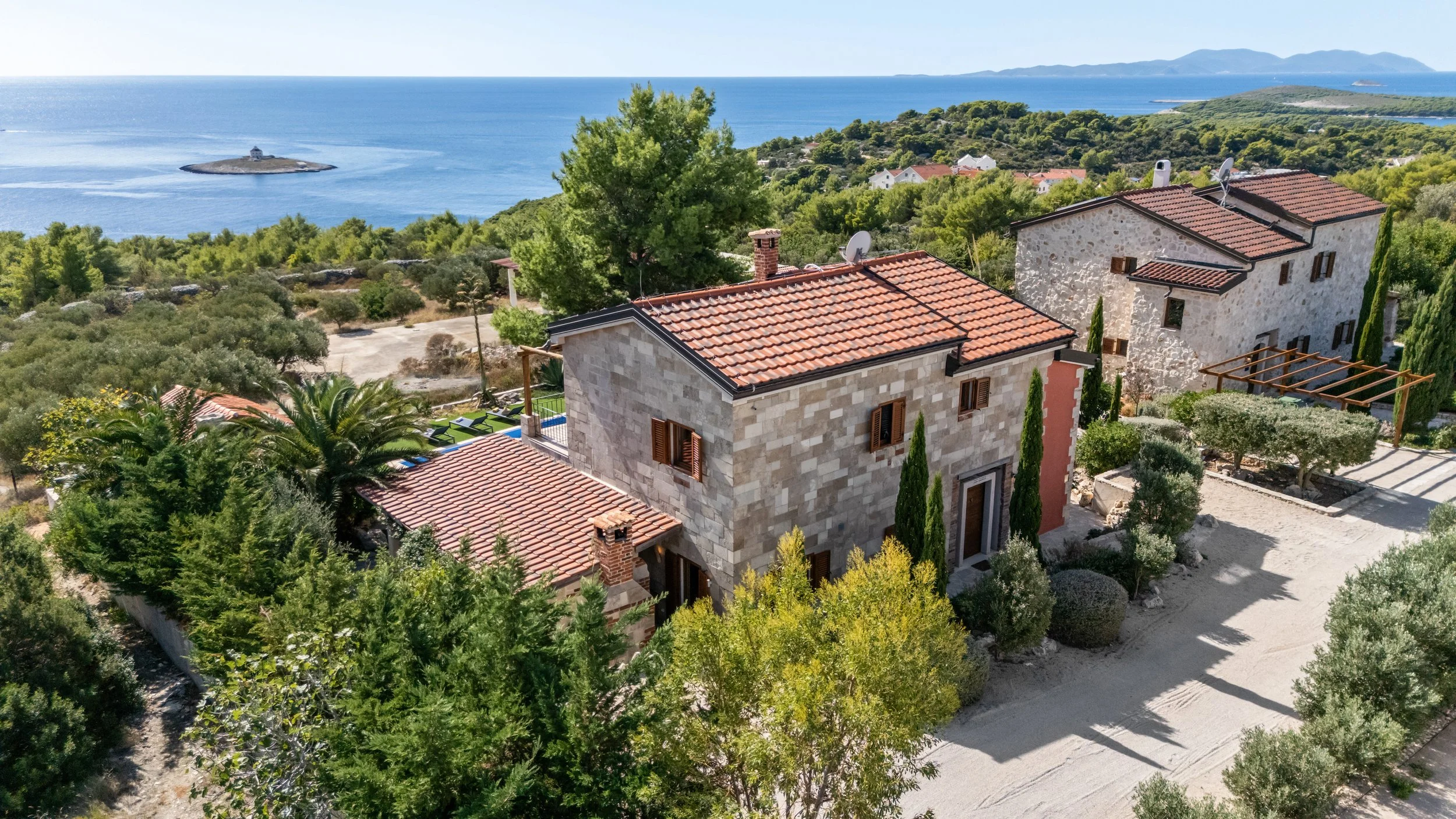 Aerial view of Mediterranean-style houses with tiled roofs surrounded by lush trees, overlooking a coastal landscape with an island and the ocean.