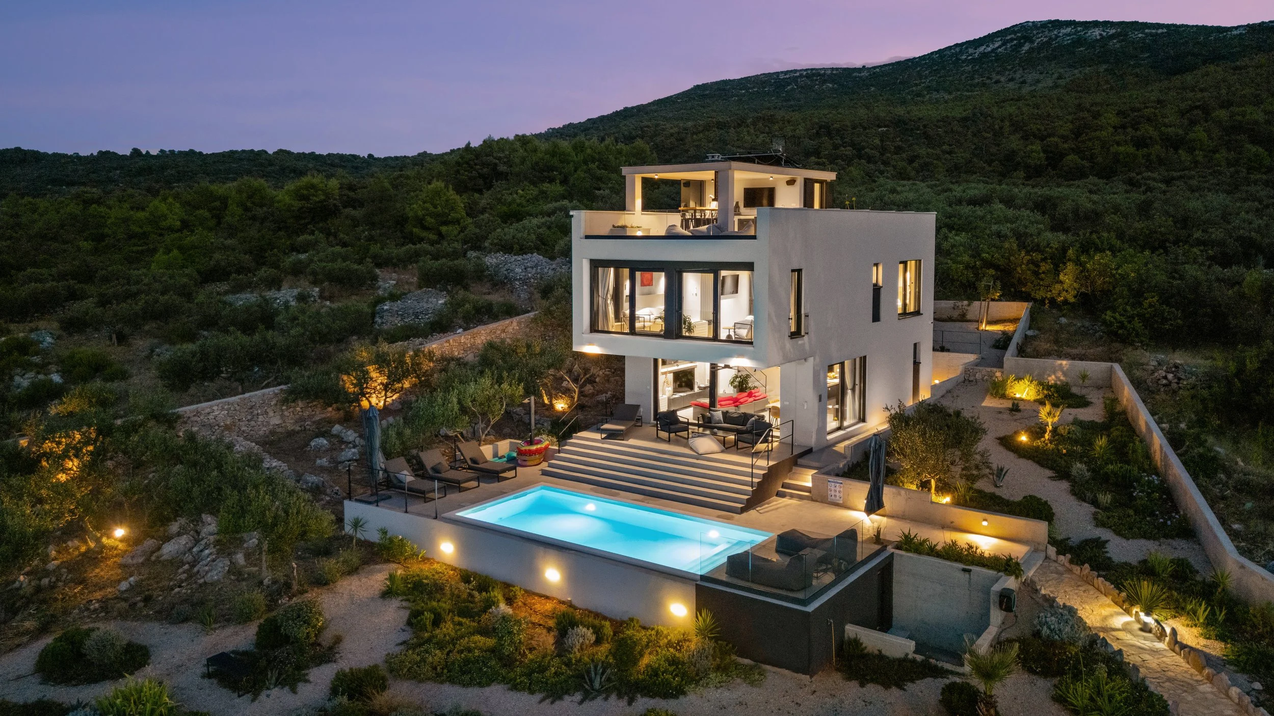 A modern multi-story house on a hillside at dusk, illuminated with outdoor lighting. The house has large windows, a balcony, and an outdoor pool with lounge chairs and patio furniture surrounding it. The landscape features desert plants and rocks.