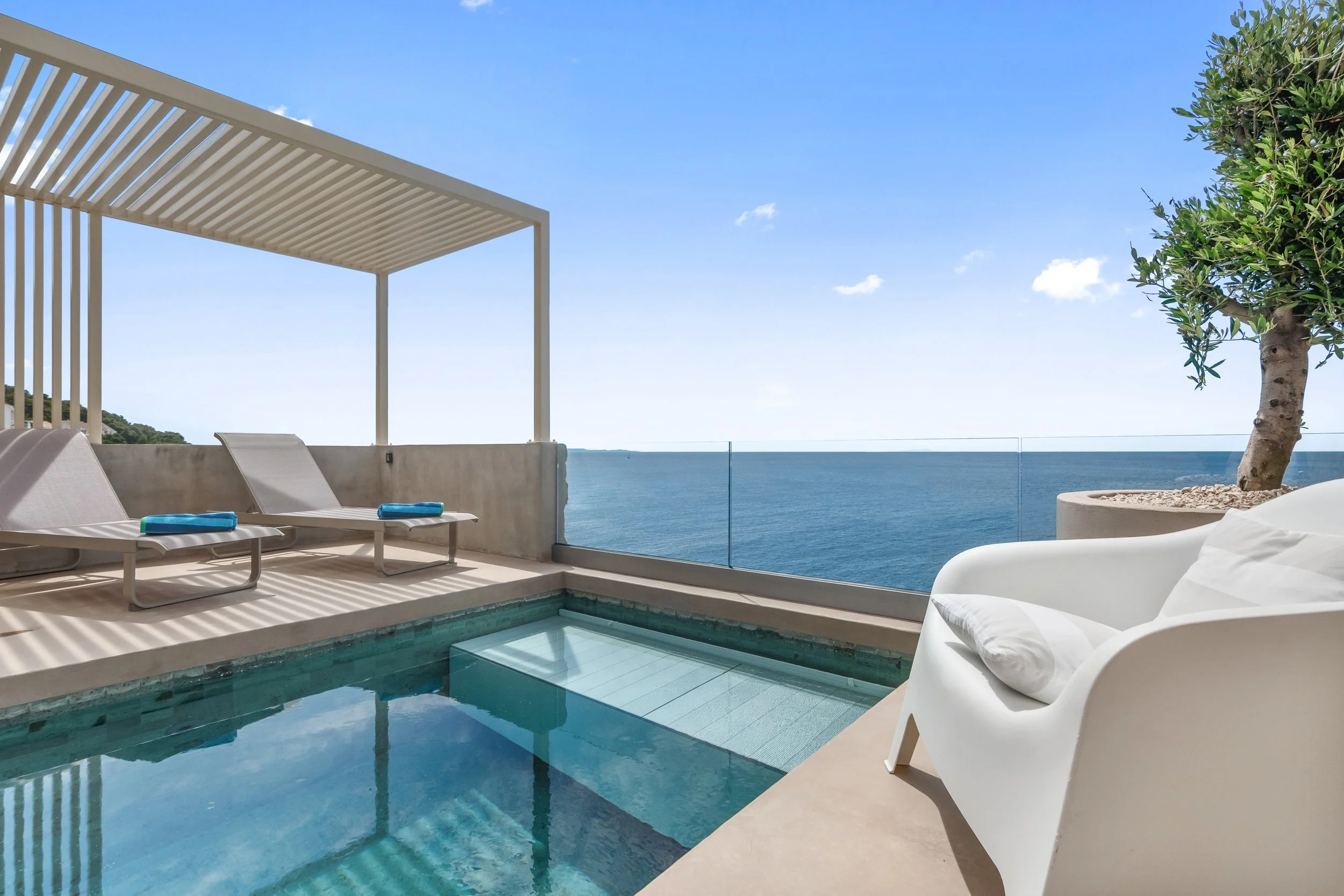 A rooftop balcony with a small pool, two lounge chairs with towels, a white lounge chair with a pillow, and a potted tree overlooking the ocean under a blue sky.