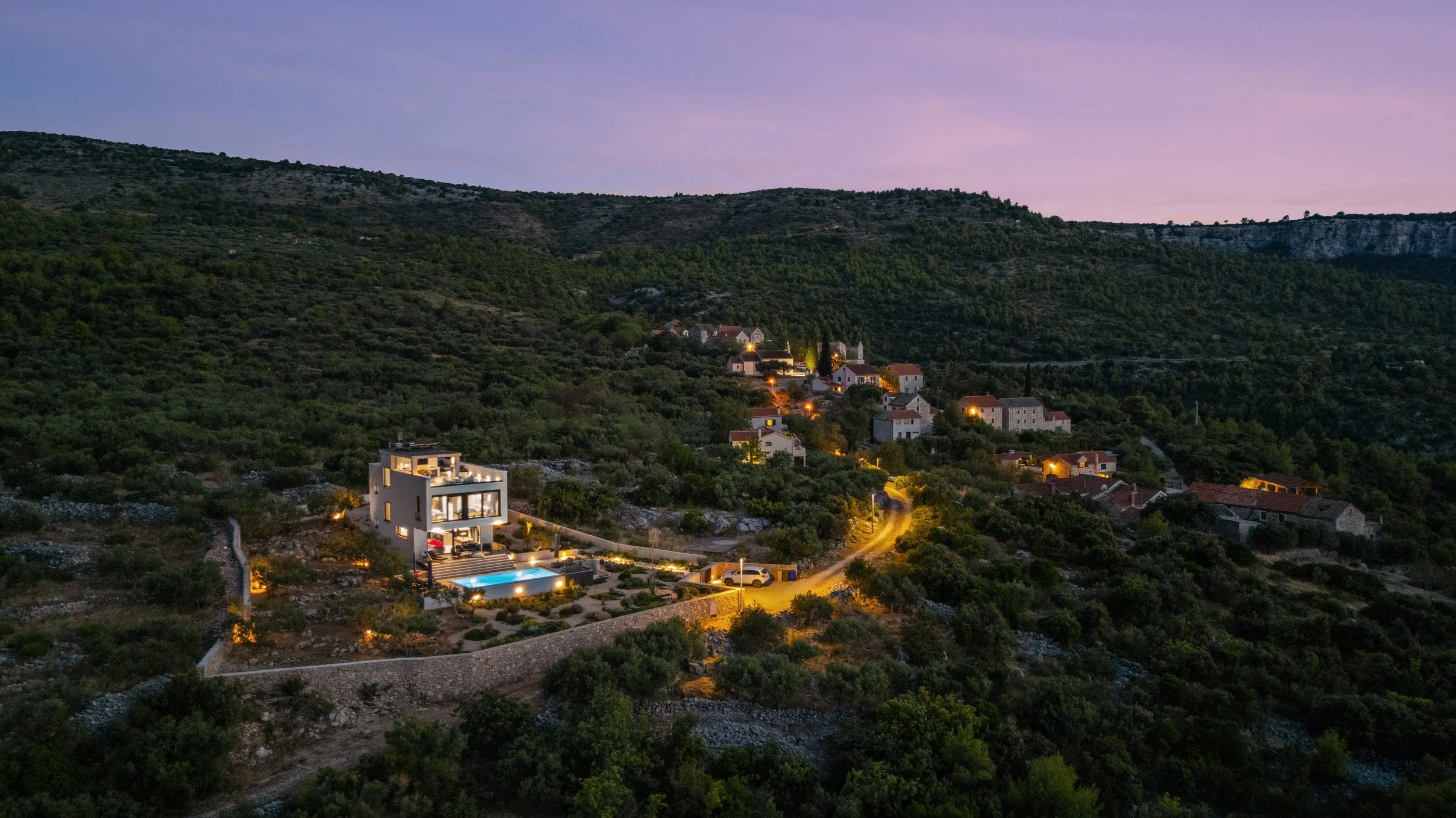 A modern house with a swimming pool situated on a hillside, surrounded by trees and other houses, during dusk with a purple sky.