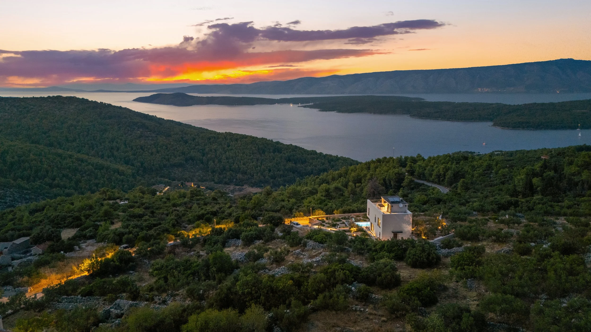 A scenic view of a sunset over a large lake, surrounded by green hills and mountains. In the foreground, a modern white house with outdoor lighting is nestled among trees and rocks.