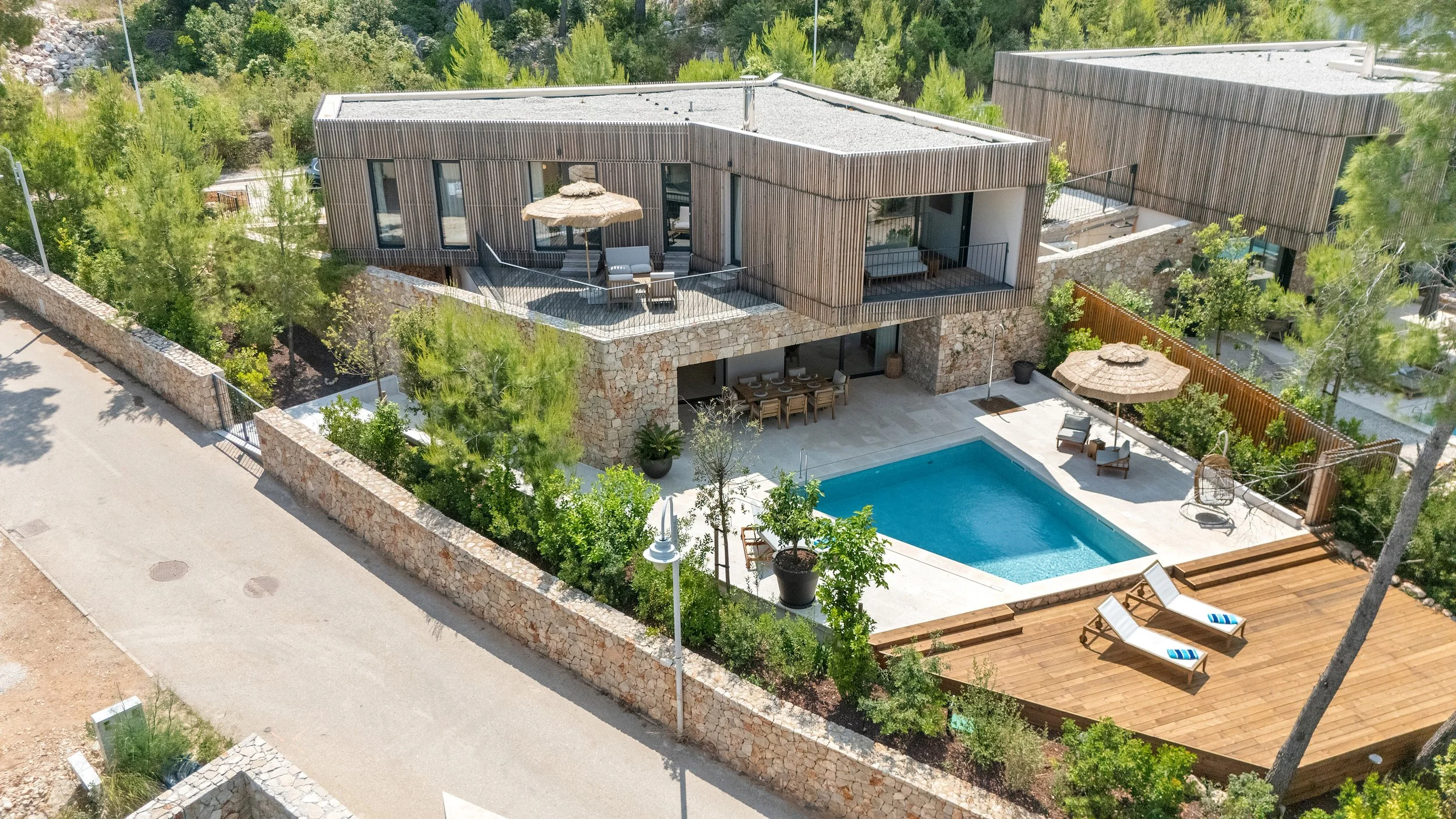 Aerial view of a modern house with a swimming pool, wooden deck, and outdoor seating area surrounded by lush greenery and a stone wall.