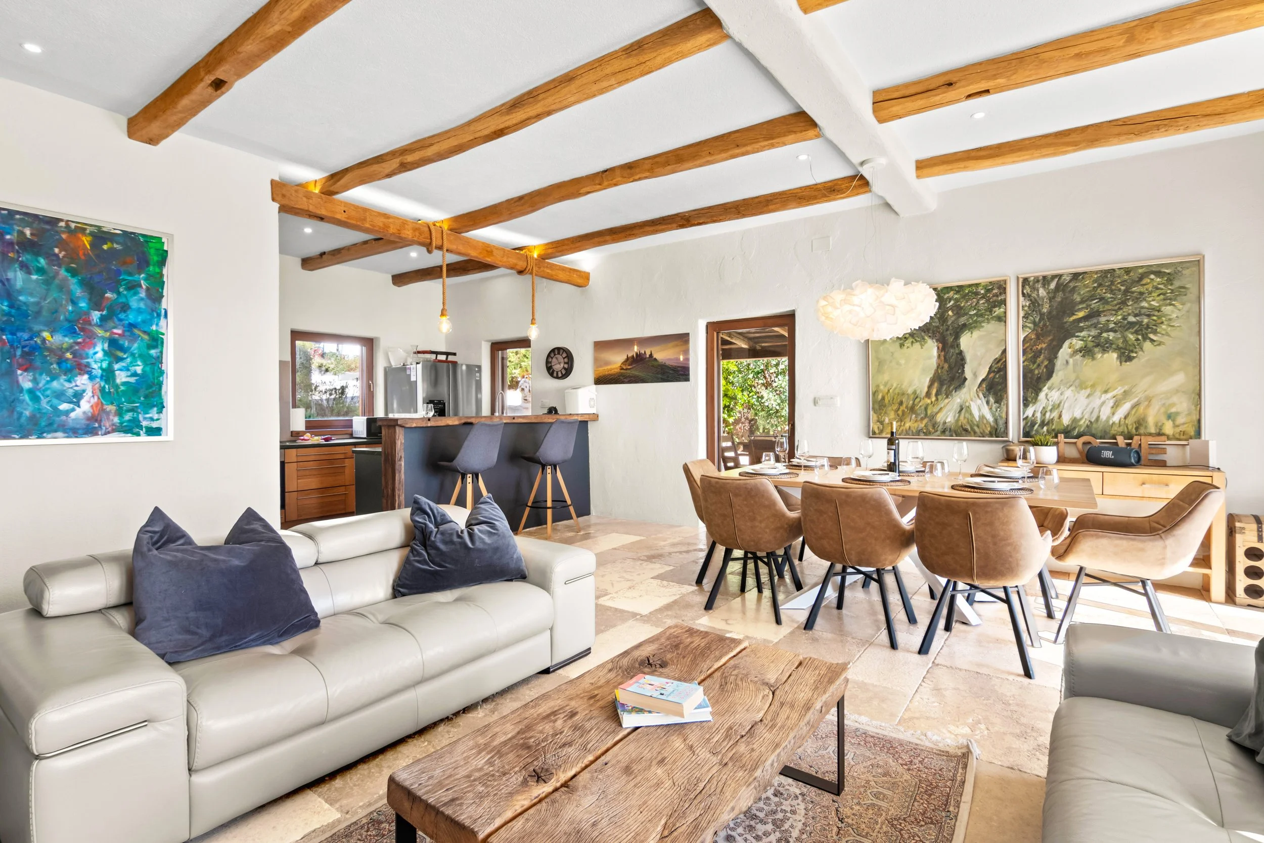 Open-concept living and dining area with white leather couches, a wooden dining table with beige chairs, artistic wall paintings, wooden ceiling beams, and a kitchen with a bar counter.
