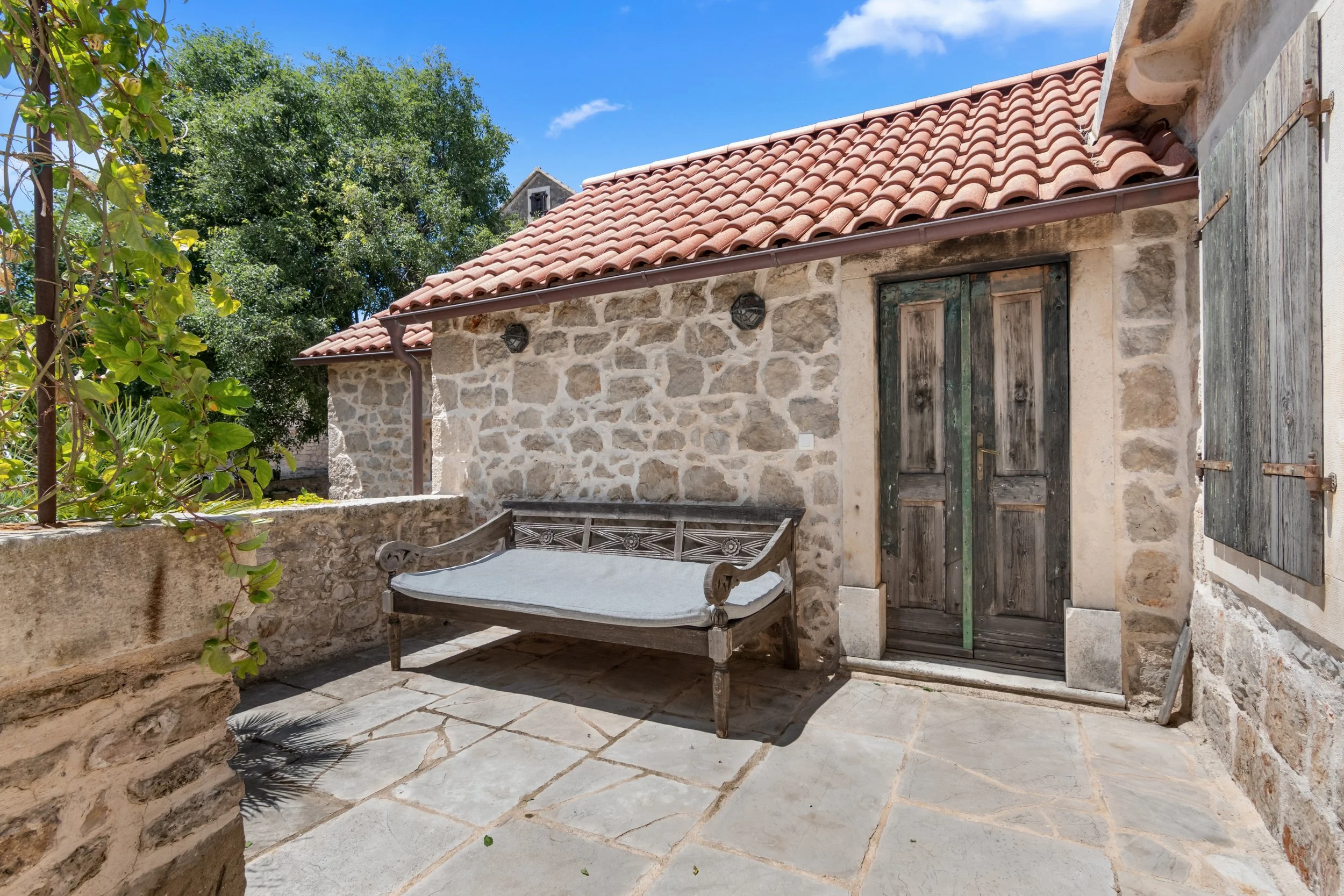 Stone house exterior with wooden door, window shutters, red tile roof, and outdoor bench on a stone patio under a blue sky.