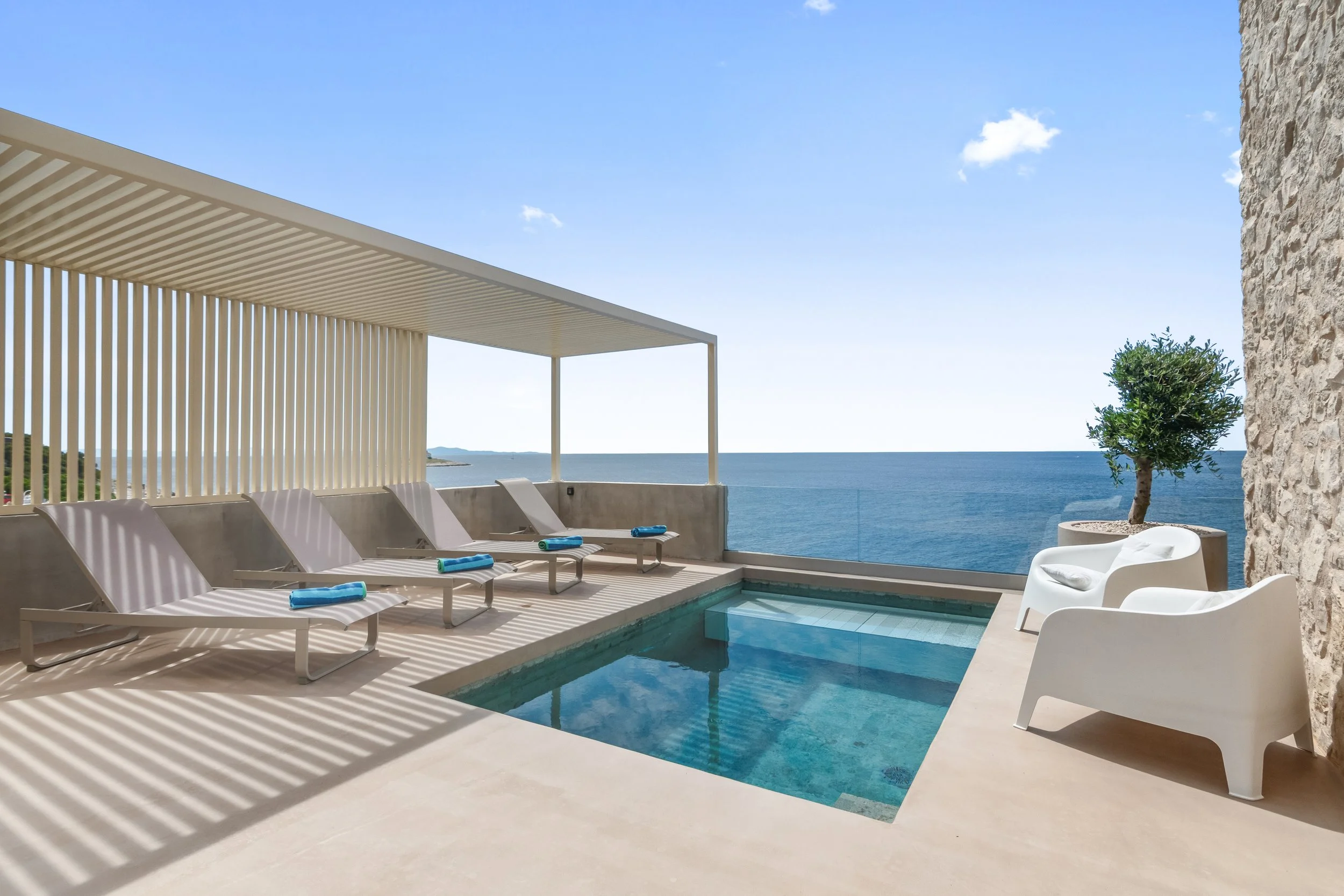 Poolside balcony with four lounge chairs, two white armchairs, and a potted tree overlooking the ocean under a partly cloudy sky.