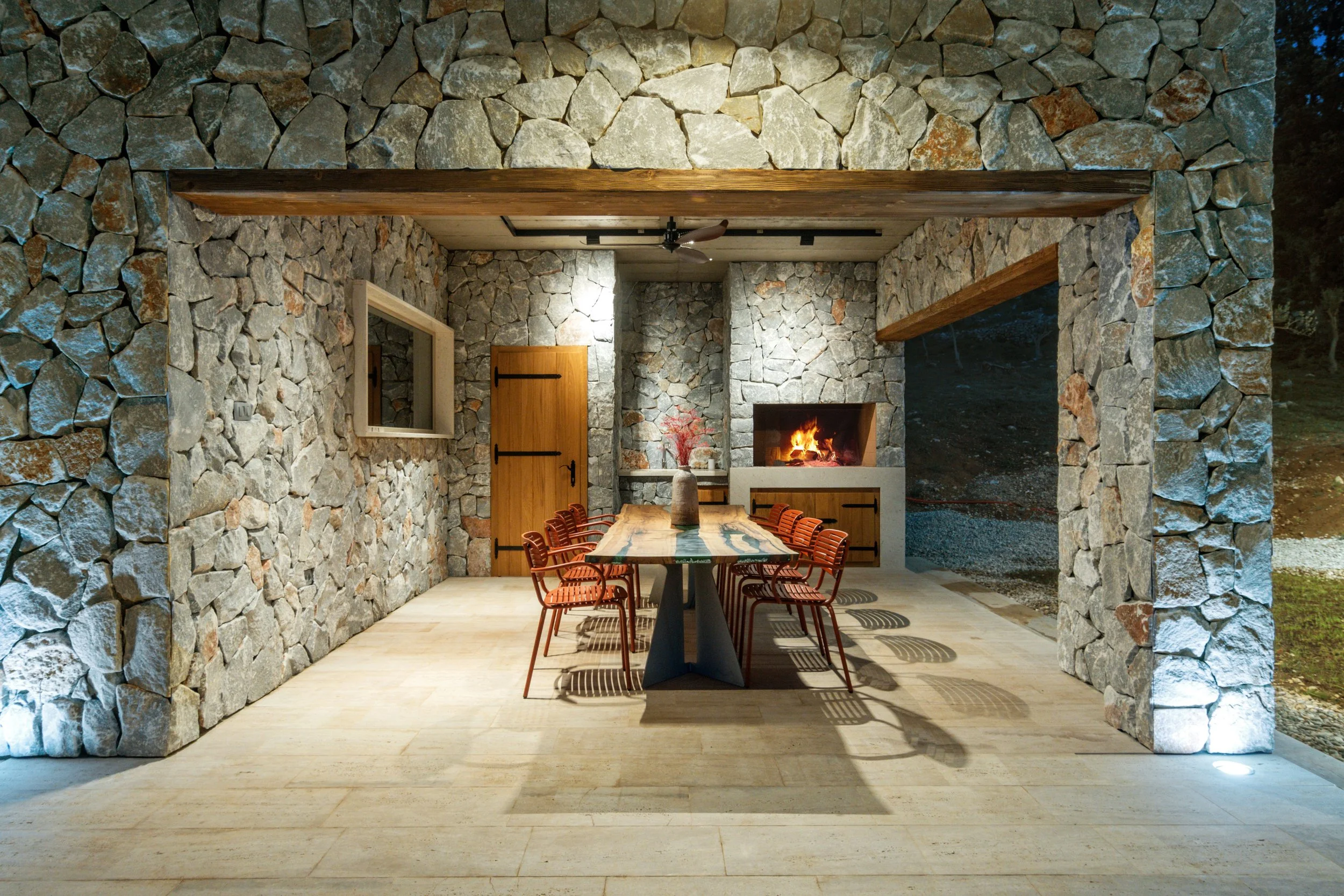 A rustic indoor dining area with stone walls, a wooden door, a long wooden dining table with wooden chairs, and a fireplace with a cozy fire.