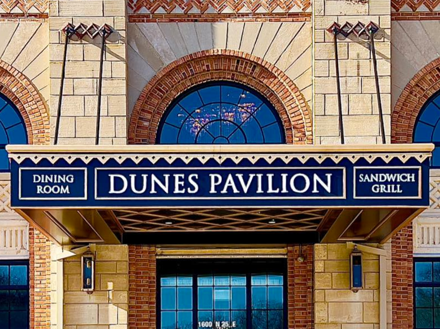 Sign for Dunes Pavilion with options for dining room, sandwich grill, and outdoor seating, in front of a brick building with arched windows.