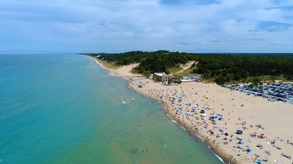 Aerial view of a crowded sandy beach with many umbrellas, near the ocean shoreline, with green trees and hills in the background.
