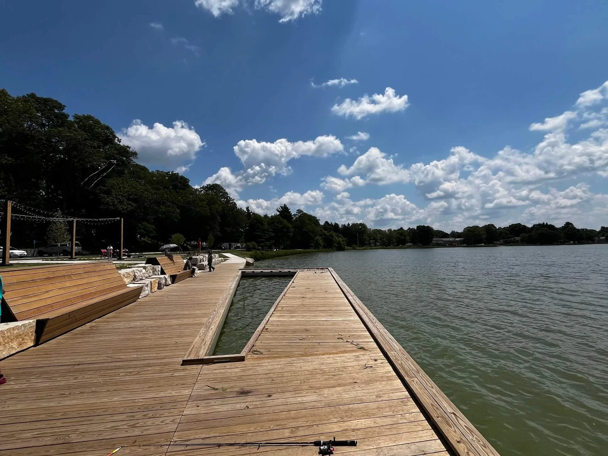 View of a wooden dock along a lake with benches, trees, and a blue sky with clouds.