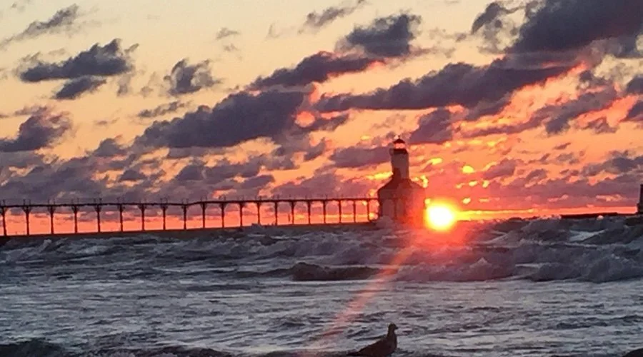 Sunset over a lighthouse and pier with waves crashing on the shore.