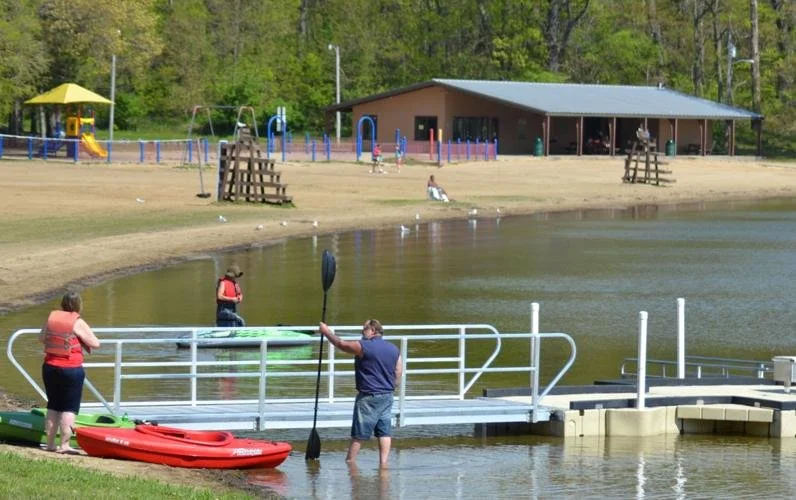 People enjoying water activities at a lake, including kayaking and paddleboarding, with a playground and a pavilion in the background.
