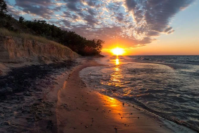 Sunset over a beach with waves gently crashing, a rocky shoreline to the left, and a partly cloudy sky.