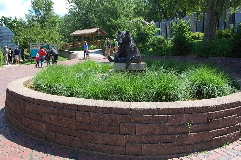 A zoo entrance with a sign that says "Welcome!" People walking toward the entrance, including children and adults. There is a bear statue sitting on a bench surrounded by green grass in a circular brick planter.