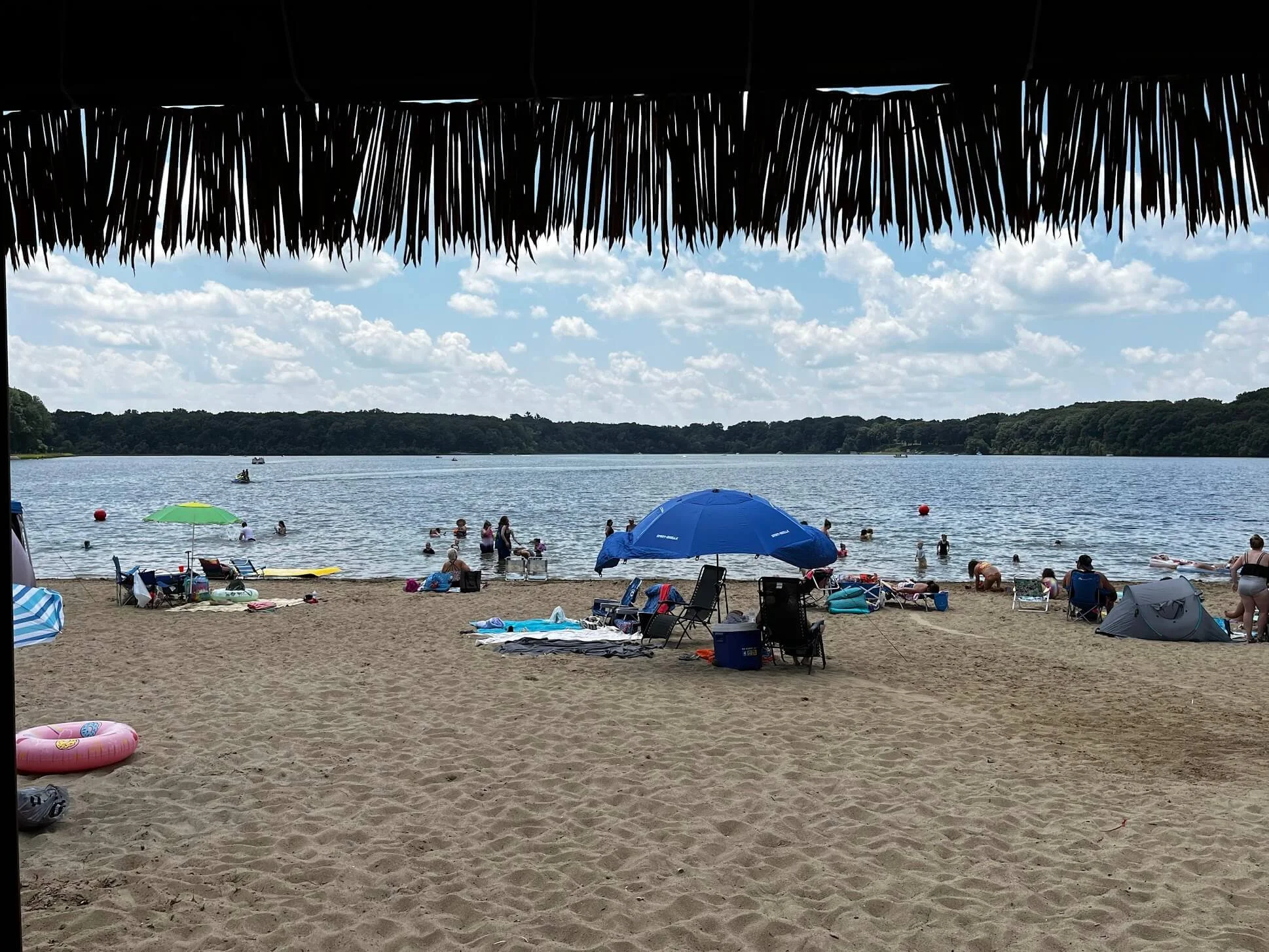 Beach scene with people swimming and relaxing. There are umbrellas, chairs, and towels on the sand, with a calm lake and trees in the background. The sky is partly cloudy.