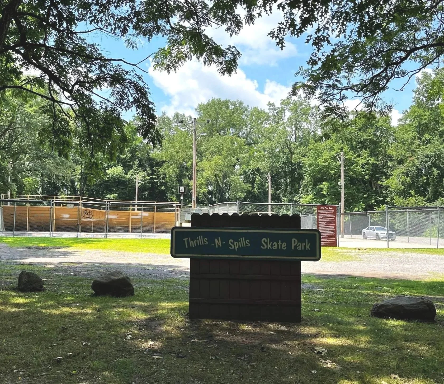 Sign at Thrills N Spills Skate Park with trees, a fence, skateboard ramp, and parked vehicle in background.