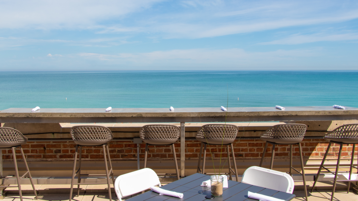 Beachside restaurant with six bar stools along a bar overlooking the ocean, a small table set with utensils and a potted plant, under a partly cloudy sky.