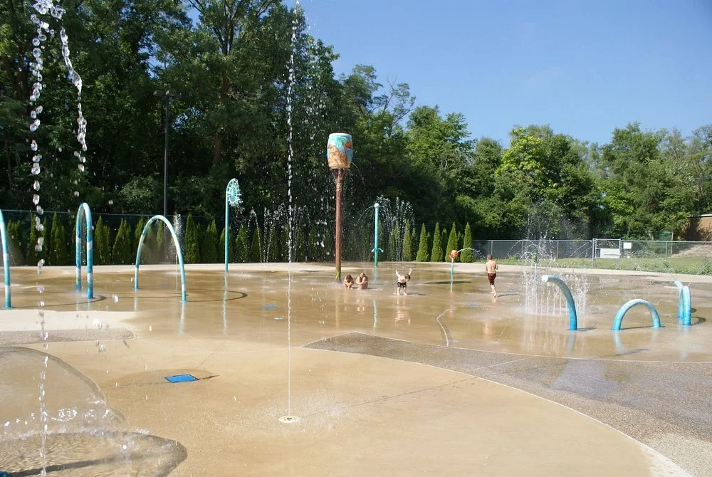 Children and adults playing in a splash pad with multiple water sprays and fountains during daytime, surrounded by green trees and a fenced area.