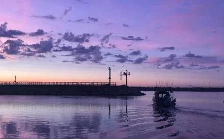 A boat in a harbor at sunset with purple and orange sky, and a pier with structures extending into the water.