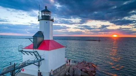 A lighthouse on a rocky shoreline during sunset, with a partly cloudy sky and calm water surrounding it.