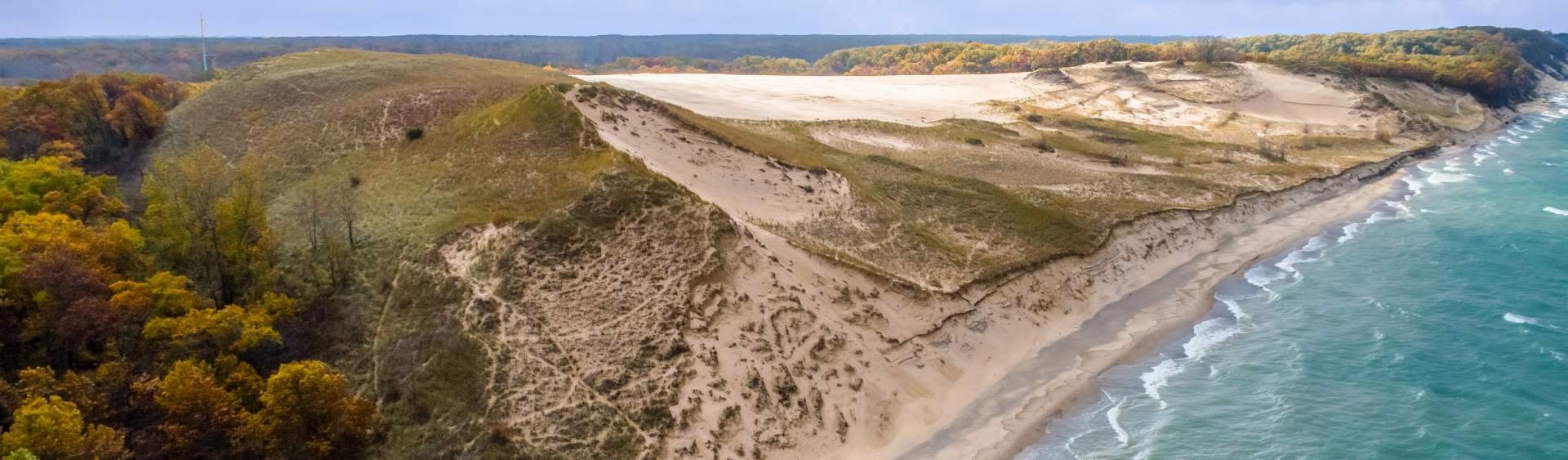 Aerial view of white sandy cliffs and beach along a blue ocean, with colorful autumn trees on the left side of the image.